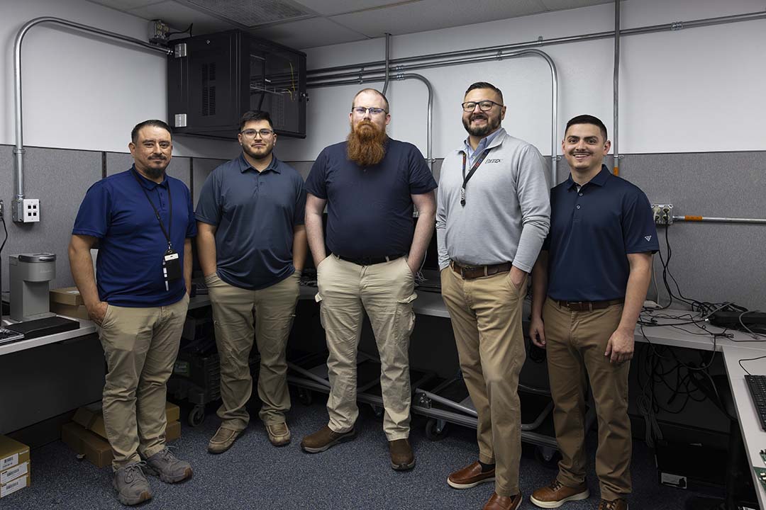 Five men stand in a row inside a large cubicle. All are wearing khakis of various shades. Three on the left and the one on the right end are wearing blue, short-sleeved shirts. The man second from the right is wearing a long-sleeved grey sweater. All have dark hair except for the man in the middle who is red-headed.