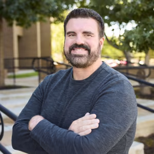 A man with dark, short-cut hair and trimmed beard is outside smiling, leaning up against the railing of steps leading up to a building. He’s wearing a dark gray shirt, arms crossed. A few trees with green leaves are in the back ground. 